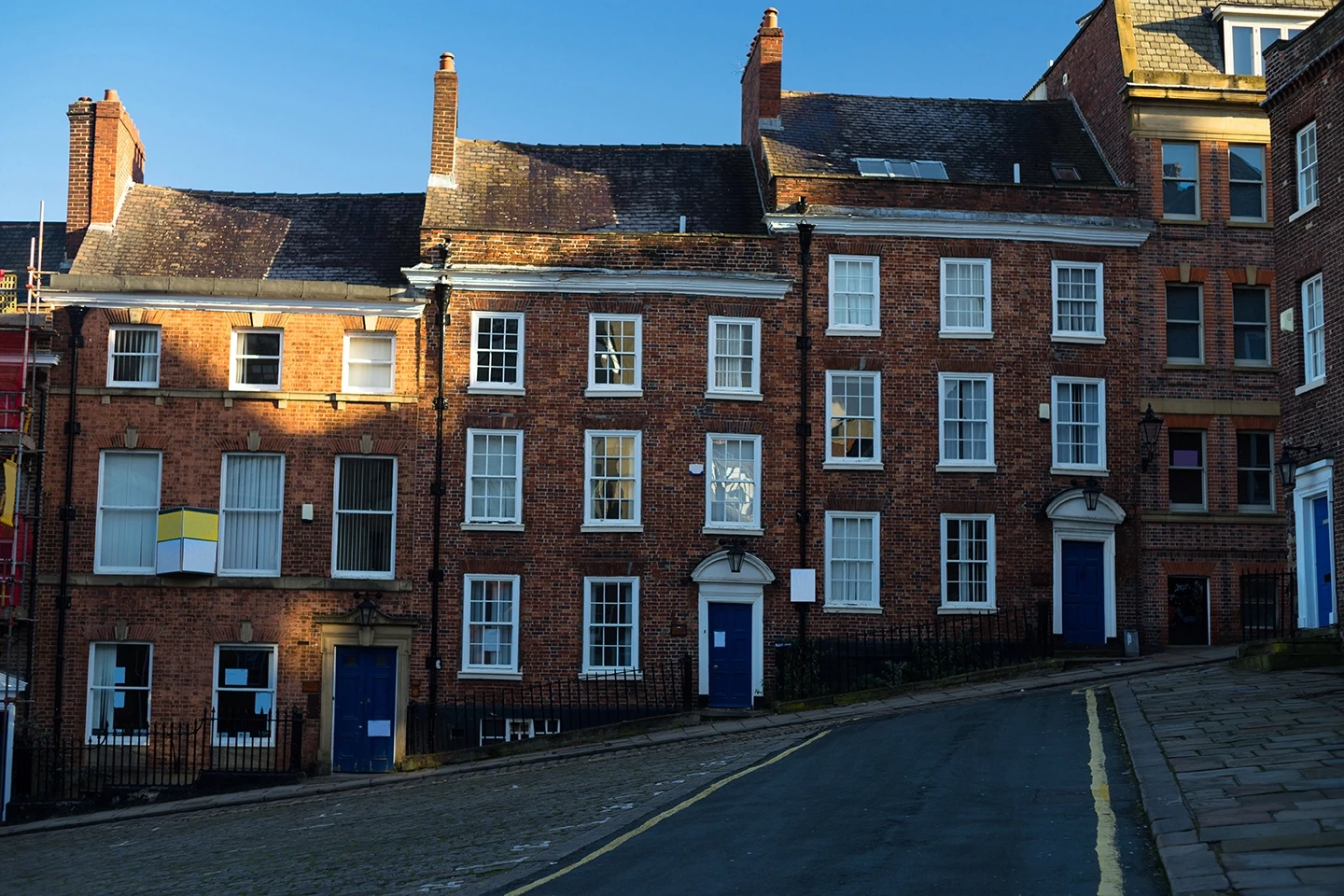 Two rows of brick buildings on a sloping street, featuring white windows and blue doors, under a clear blue sky.