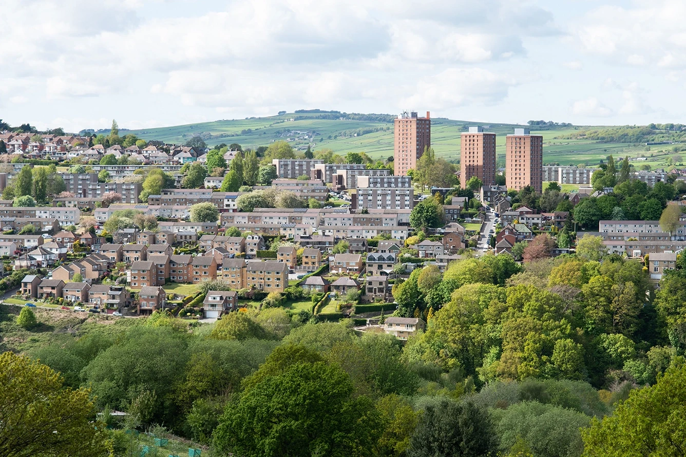 A panoramic view of a residential area with various houses and tall buildings, surrounded by green hills and trees under a partly cloudy sky.