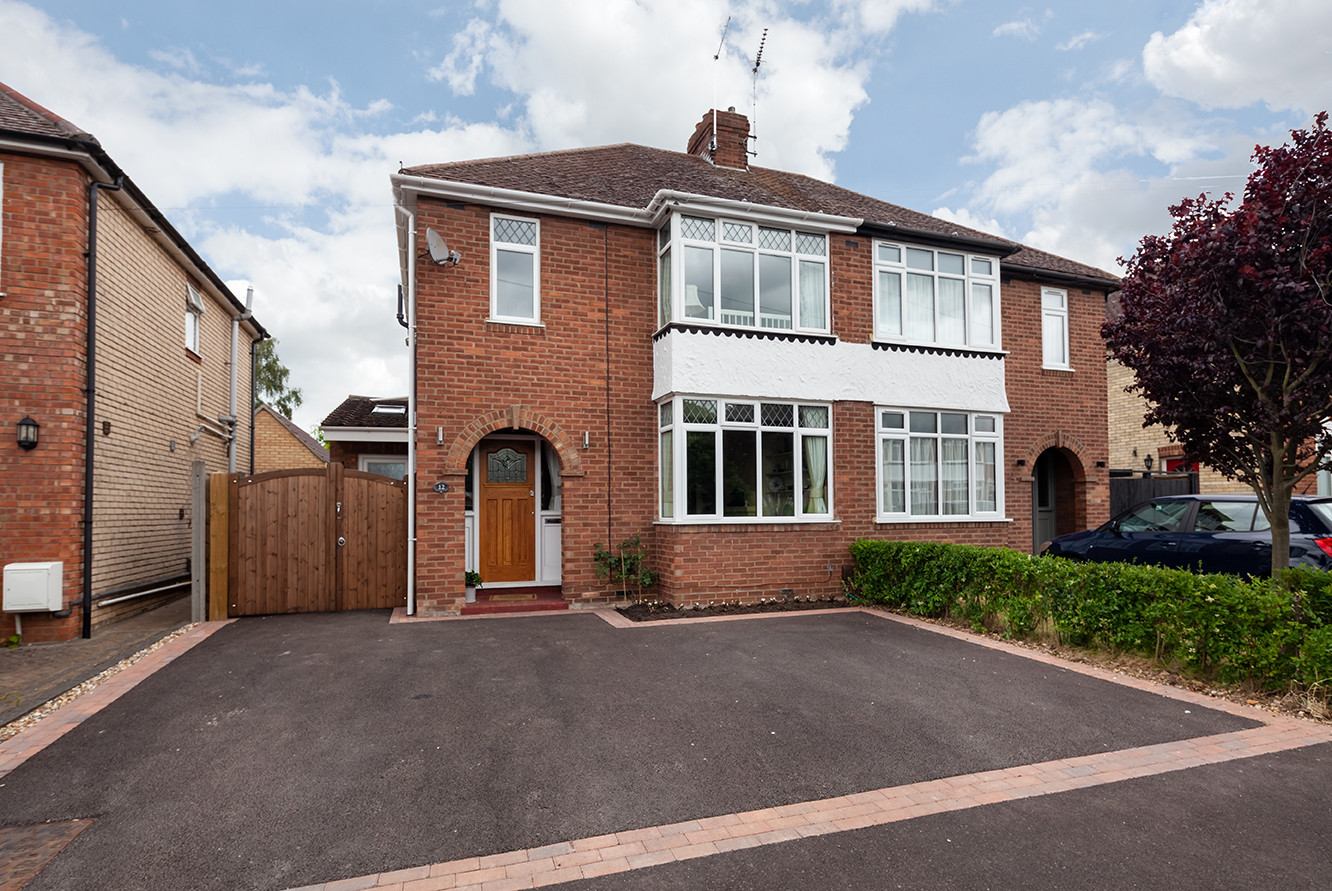 A charming red-brick semi-detached house with a wooden front door, surrounded by well-maintained gardens and a paved driveway.